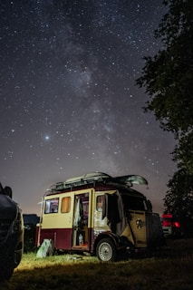 A vintage camper van is parked under a sky full of stars, with the Milky Way clearly visible. The van is illuminated softly, suggesting it is nighttime, and is surrounded by other vehicles and trees. Inside, the van appears to be lived in, with clothes and other belongings visible.