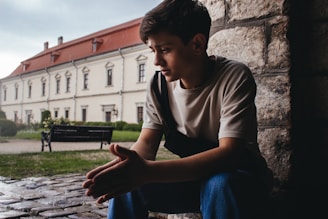 A young person is seated near the entrance of a stone building, appearing to be deep in thought. In the background, there is an old, large building with a red roof and multiple windows, typical of historical architecture. The setting appears to be an outdoor area with a park bench and some greenery.