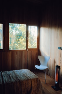 A cozy room with wooden walls and flooring is gently illuminated by natural light coming through a window. There is a modern white chair placed against the wall and a small electric heater nearby. A bed with a textured blanket is partially visible in the foreground.