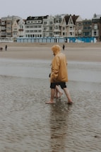 A person in a yellow raincoat and black shorts walks barefoot along a wet and sandy beach. Behind them, a row of older, diverse architectural-style buildings stands alongside the coast. The tide is out, leaving puddles on the sand, and the atmosphere appears overcast.