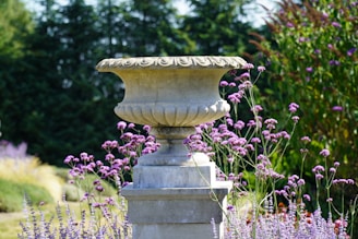 A large and ornate stone urn is surrounded by vibrant purple flowers and lush greenery. The urn is positioned on a pedestal and is intricately detailed with carvings. In the background, there are dense, dark green trees creating a natural backdrop.