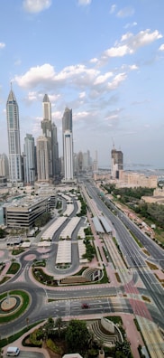 An urban skyline featuring a collection of tall, modern skyscrapers set against a background of scattered clouds and blue sky. A wide multi-lane highway runs through the cityscape, with minimal traffic visible. The surrounding area includes well-maintained roads and green spaces. The architectural design includes some curved elements and a mix of glass and concrete facades.