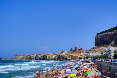 A vibrant beach scene with crowded sandy shores. Numerous umbrellas in various colors provide shade for beachgoers. People are enjoying the sea, with some swimming and others sunbathing. A rocky cliff with dense greenery and historic buildings, featuring red-tiled roofs, dominate the background. The townscape extends along the coastline under a clear blue sky.