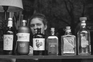 A black and white image displaying a collection of various gin bottles lined up on a surface, with distinctive and artistic labels. Behind the bottles, there is a person looking forward, partially obscured. The backdrop appears to be a dimly lit room with dark furnishings and a lamp above the bottles providing some illumination.