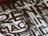 A close-up view of a section of a stone or metal engraving featuring large, bold letters intertwined with classical architectural elements. The surface appears textured, adding depth and contrast.