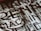 A close-up view of a section of a stone or metal engraving featuring large, bold letters intertwined with classical architectural elements. The surface appears textured, adding depth and contrast.