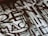 A close-up view of a section of a stone or metal engraving featuring large, bold letters intertwined with classical architectural elements. The surface appears textured, adding depth and contrast.