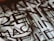 A close-up view of a section of a stone or metal engraving featuring large, bold letters intertwined with classical architectural elements. The surface appears textured, adding depth and contrast.