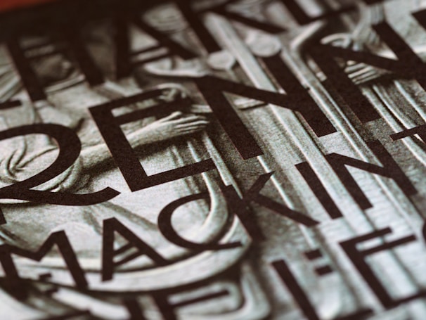 A close-up view of a section of a stone or metal engraving featuring large, bold letters intertwined with classical architectural elements. The surface appears textured, adding depth and contrast.