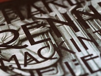 A close-up view of a section of a stone or metal engraving featuring large, bold letters intertwined with classical architectural elements. The surface appears textured, adding depth and contrast.