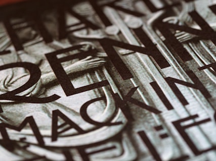 A close-up view of a section of a stone or metal engraving featuring large, bold letters intertwined with classical architectural elements. The surface appears textured, adding depth and contrast.