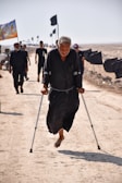 A man wearing dark clothing walks on a sandy path using crutches. He is barefoot and is surrounded by people and flags on a barren landscape.