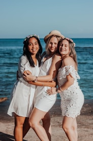 Three women are joyfully embracing each other on a beach with a backdrop of the ocean. They are wearing white dresses and floral accessories and appear to be enjoying a sunny day.