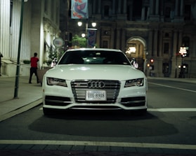 A white Audi car is parked on an urban street at night, with Virginia license plates visible. The background consists of grand architectural buildings illuminated by street lights, adding a sense of elegance to the scene. A person wearing a red shirt can be seen walking on the sidewalk.