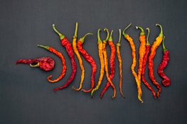 A collection of dried, colorful chili peppers is arranged on a dark, textured surface. The peppers range in color from vibrant red to deep orange and have a wrinkled appearance. Some of the stems are still attached, giving a natural, organic look.