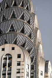 An architectural detail of a skyscraper featuring art deco design elements, characterized by its geometric patterns and metallic surface. The building's facade is decorated with triangular motifs and has a distinctly vintage appearance, highlighting its intricate craftsmanship.