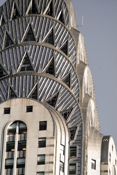 An architectural detail of a skyscraper featuring art deco design elements, characterized by its geometric patterns and metallic surface. The building's facade is decorated with triangular motifs and has a distinctly vintage appearance, highlighting its intricate craftsmanship.