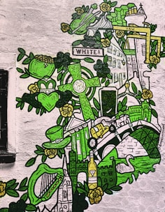 A vibrant mural featuring various Irish symbols and landmarks. The artwork includes a Celtic cross, leprechaun hat, harp, swan, and the Harland & Wolff cranes, all painted in shades of green and yellow. The imagery is intricately detailed with floral motifs and bubbles scattered throughout the piece.