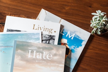 A collection of magazines is fanned out on a wooden table, featuring covers with titles such as 'Living' and 'Hale for KO OLINA'. The cover images include a beach scene and a person in a hat, with a small succulent plant visible to the right.