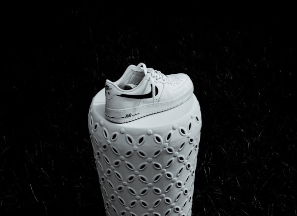 A white sneaker with a black swoosh logo sits atop a decorative ceramic pedestal. The pedestal features an intricate pattern of oval cut-outs, providing an ornate contrast to the simple design of the sneaker. The background is dark, emphasizing the shoe and pedestal.