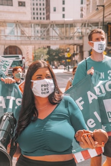 A group of people participating in a protest in an urban setting. They are wearing face masks with 'Pro Life Dem' written on them. One person is holding a small baby doll and a megaphone, while others hold a large banner.