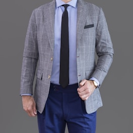 A person is wearing a grey checkered suit jacket over a light blue shirt paired with a black tie and dark blue trousers. The individual's left hand is adjusting the jacket, and they are wearing a watch on their left wrist.