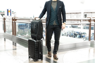 man in blue suit jacket and black pants standing beside black luggage bag