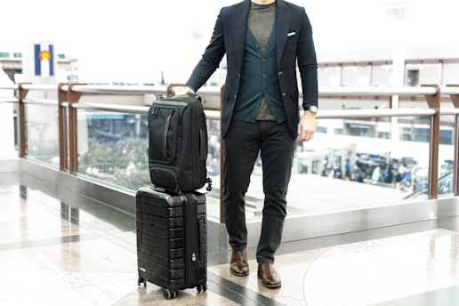 man in blue suit jacket and black pants standing beside black luggage bag
