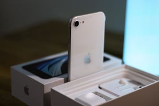 A white smartphone with an Apple logo is partially removed from its box, showcasing the back of the phone. The box is placed on a wooden surface with the phone standing upright. The internal compartments of the box are visible, including the designated slots for accessories.