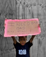 A child stands holding a handwritten cardboard sign with the message 'I need my dad. It's time 4 change.' The child is wearing a T-shirt with the words 'Black Lives Matter' against a backdrop of wooden boards.