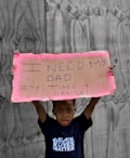 A child stands holding a handwritten cardboard sign with the message 'I need my dad. It's time 4 change.' The child is wearing a T-shirt with the words 'Black Lives Matter' against a backdrop of wooden boards.
