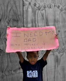 A child stands holding a handwritten cardboard sign with the message 'I need my dad. It's time 4 change.' The child is wearing a T-shirt with the words 'Black Lives Matter' against a backdrop of wooden boards.