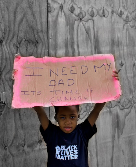 A child stands holding a handwritten cardboard sign with the message 'I need my dad. It's time 4 change.' The child is wearing a T-shirt with the words 'Black Lives Matter' against a backdrop of wooden boards.