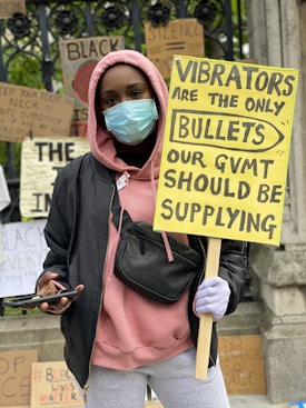 A person wearing a mask holds a large yellow protest sign with bold lettering. They are dressed in a black jacket and pink hoodie, carrying a small black bag across their chest. Several other protest signs with various messages are visible in the background.