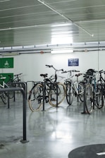 Several bicycles are parked in an indoor bicycle parking area with metal racks. The room has a polished concrete floor and a ceiling with tube lighting. There are signs on the wall related to cycling.
