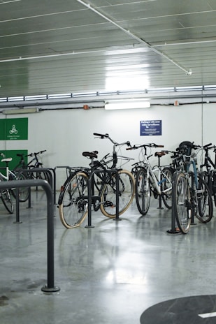Several bicycles are parked in an indoor bicycle parking area with metal racks. The room has a polished concrete floor and a ceiling with tube lighting. There are signs on the wall related to cycling.
