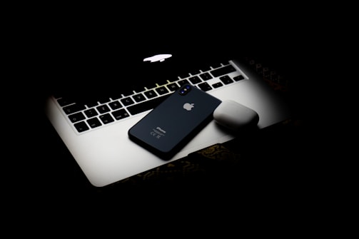 A sleek Apple laptop with its logo glowing subtly is partially covered by an iPhone and a pair of wireless earbuds. The objects are arranged on a dark surface, creating a modern and minimalistic composition with a focus on technology.