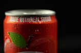 A close-up of a red soda can with droplets of water on its surface. The top of the can is visible, with the phrase 'MADE WITH REAL SUGAR' printed in white letters near the rim. Part of a green leaf is also visible on the can's design.