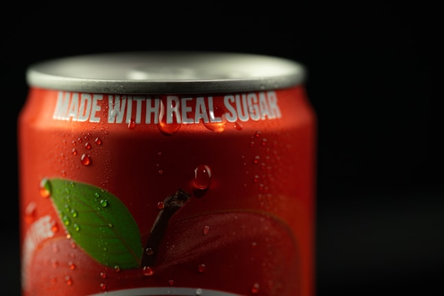 A close-up of a red soda can with droplets of water on its surface. The top of the can is visible, with the phrase 'MADE WITH REAL SUGAR' printed in white letters near the rim. Part of a green leaf is also visible on the can's design.