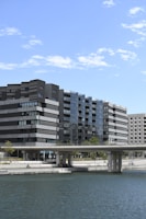Modern architectural buildings with a sleek design line a waterfront. The structures feature dark, rectangular patterns with glass surfaces reflecting the sky. A bridge extends across the water below, and a few people are visible in a relaxed setting near the water's edge. Trees and greenery are scattered around the walkway.