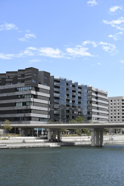 Modern architectural buildings with a sleek design line a waterfront. The structures feature dark, rectangular patterns with glass surfaces reflecting the sky. A bridge extends across the water below, and a few people are visible in a relaxed setting near the water's edge. Trees and greenery are scattered around the walkway.