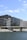 Modern architectural buildings with a sleek design line a waterfront. The structures feature dark, rectangular patterns with glass surfaces reflecting the sky. A bridge extends across the water below, and a few people are visible in a relaxed setting near the water's edge. Trees and greenery are scattered around the walkway.