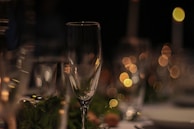 A close-up of a champagne flute surrounded by other clear glassware. The glass is set on a table with a blurred background of circular bokeh lights in warm tones, suggesting a festive or elegant atmosphere.