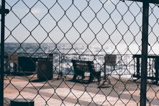 A chain-link fence partially obstructs the view of a waterfront scene with benches facing a body of water. The sunlight reflects off the water, creating a sparkling effect. A boardwalk or pier provides the setting for the benches.