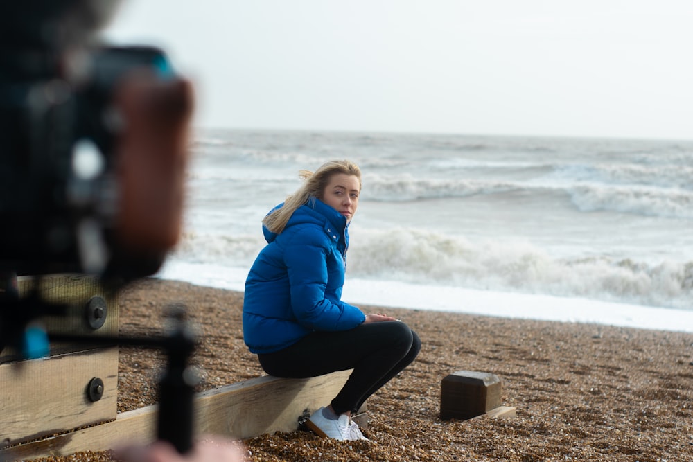 woman in blue jacket sitting on brown wooden fence near sea during daytime