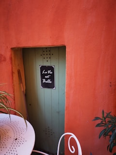 A rustic scene with a vibrant orange textured wall and a narrow green door featuring a small black sign with white script reading 'La Vie est Belle'. To the left, a white perforated metal table with greenery on it and a white curved chair add decorative elements.