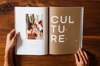 A person is holding a large, open magazine on a wooden surface. The left page features a photo of a woman holding a child in a natural, outdoor setting. Below the photo, there is a quote about love and family. The right page displays the word 'CULTURE' in large letters with smaller text around it.