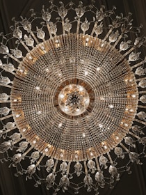 An ornate chandelier with intricate crystal and gold detailing hangs from the ceiling, radiating light. The symmetrical design includes numerous crystal elements that create an elegant and luxurious appearance.