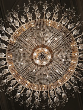 An ornate chandelier with intricate crystal and gold detailing hangs from the ceiling, radiating light. The symmetrical design includes numerous crystal elements that create an elegant and luxurious appearance.