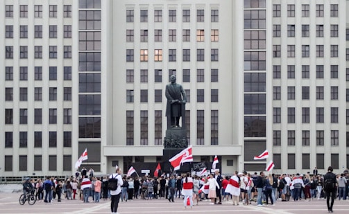 A large group of people gathers in front of a tall, rectangular government building with numerous windows. A statue stands prominently in front of the building, and many people in the crowd are holding red and white flags.